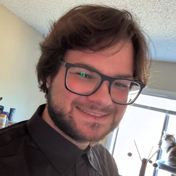 A smiling person with medium-length brown hair and glasses, wearing a dark collared shirt, with a cat in the background.