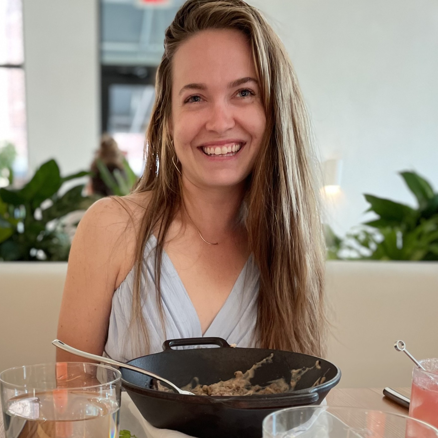 A smiling woman with long brown hair wearing a light blue top, sitting at a table with food in a restaurant.
