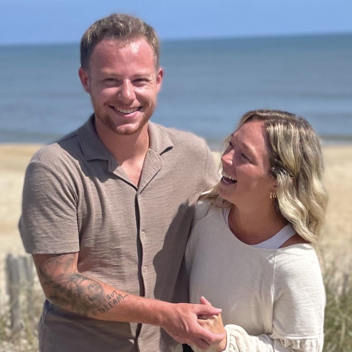 A smiling couple holds hands and laughs together while standing on a sandy beach by the ocean.