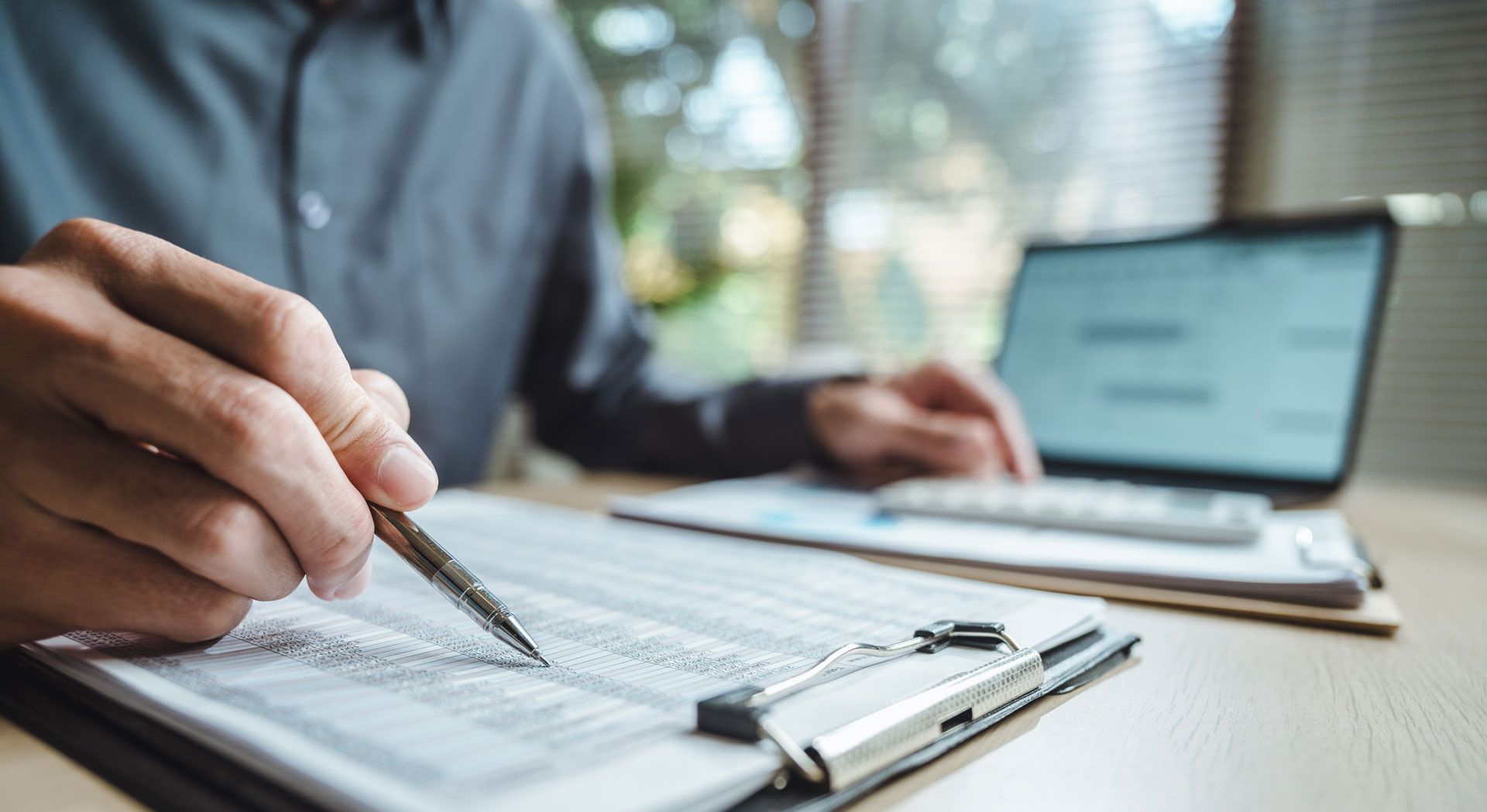 Person reviewing paperwork with a pen, a laptop and a calculator on a desk.