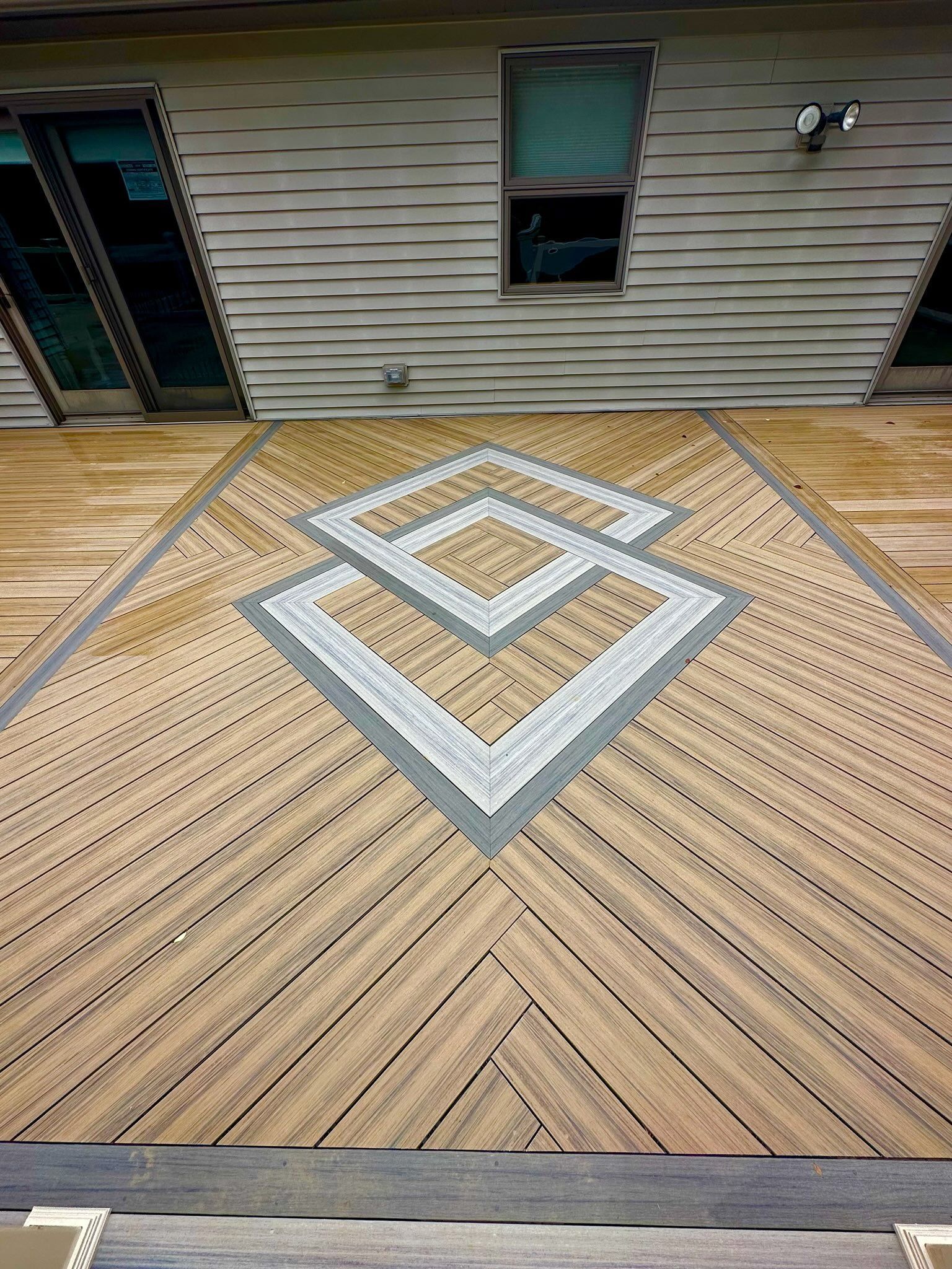 Wooden deck with a gray geometric inlay beside a house wall and windows