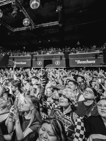 Concert crowd: people in a venue, arms raised, looking up towards stage. Disco balls hang from ceiling.