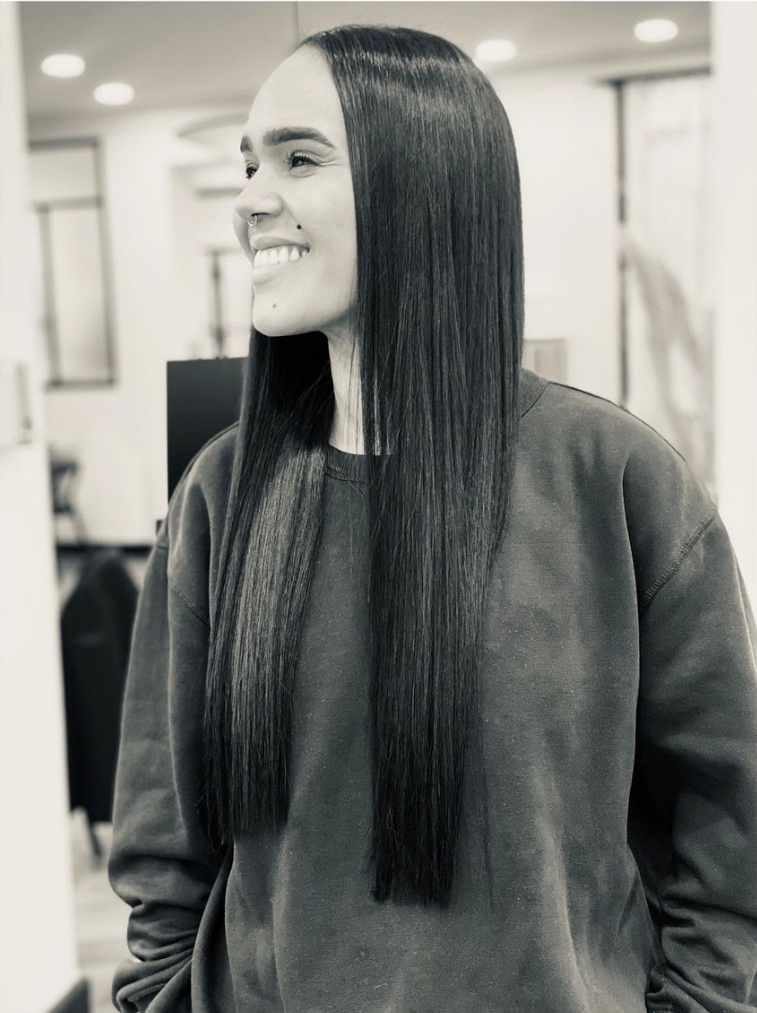 Une femme aux cheveux raides et foncés sourit ; elle porte un pull gris. Dans un salon bien éclairé.