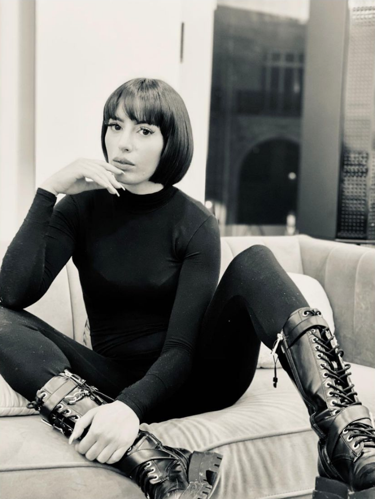 Femme vêtue de noir et portant des bottes, assise sur un canapé, le menton appuyé sur la main.