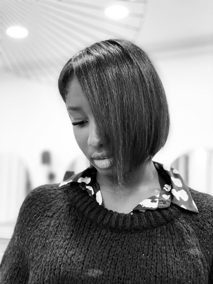 Femme aux cheveux courts et raides, regardant vers le bas. Elle porte un pull et une chemise à col. Noir et blanc.