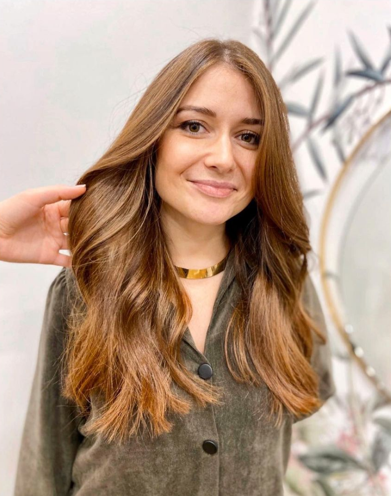 Une femme aux longs cheveux bruns ondulés, portant un ras-de-cou en or, sourit dans un salon de coiffure.