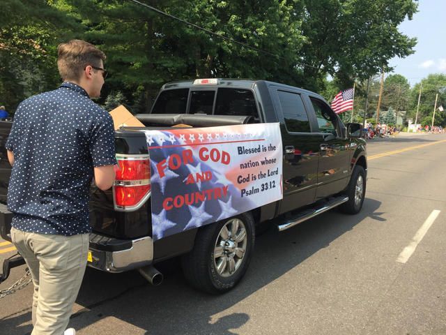 Man holding a “No God and Country” protest sign on a black pickup truck by a roadside.