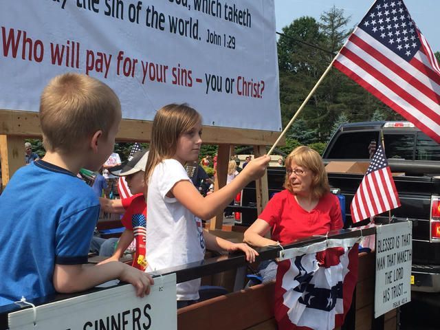 Children and a woman with protest signs and small flags at a roadside rally under a U.S. flag