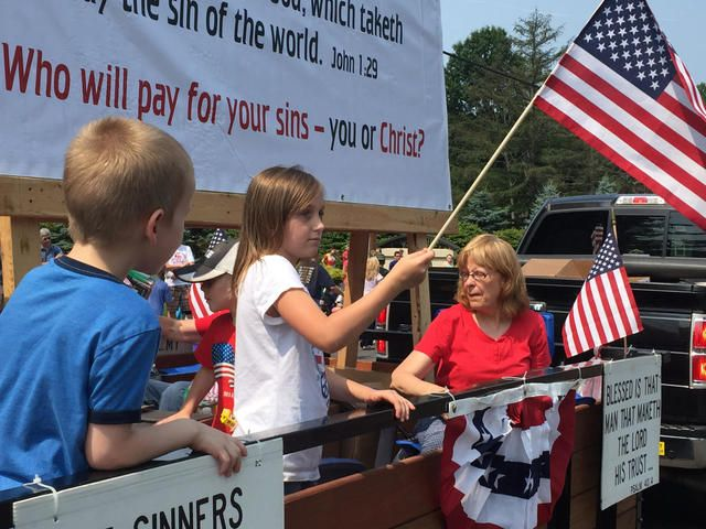 Children and a woman at a political parade with U.S. flags and a sign reading “Who will pay for your sins?”