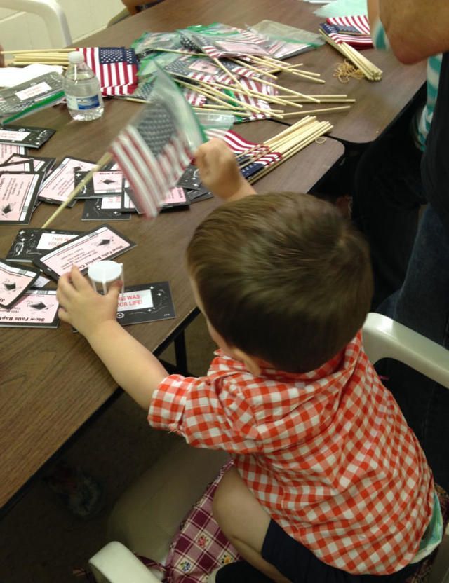 Child in a red plaid shirt at a table, assembling a small American flag craft with colorful straws.