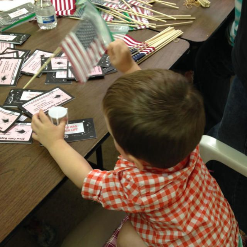 Child at a craft table waving a small American flag beside scattered flags and name cards
