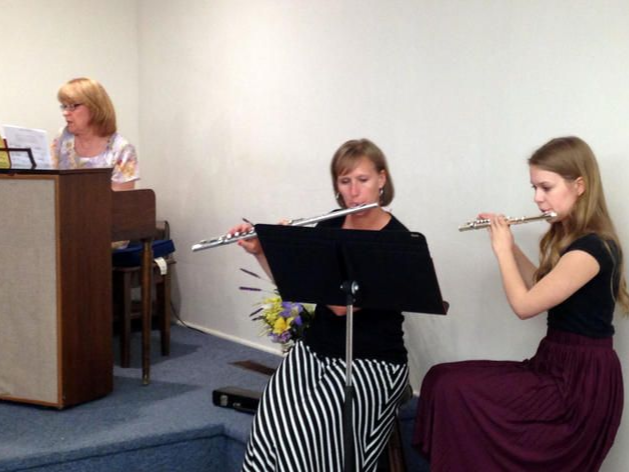 Three musicians perform in a room, with a pianist at left and two women playing flute and clarinet.