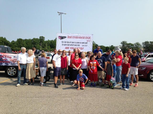 People holding a banner and flags gathered beside cars in a parking lot, posing for a group photo