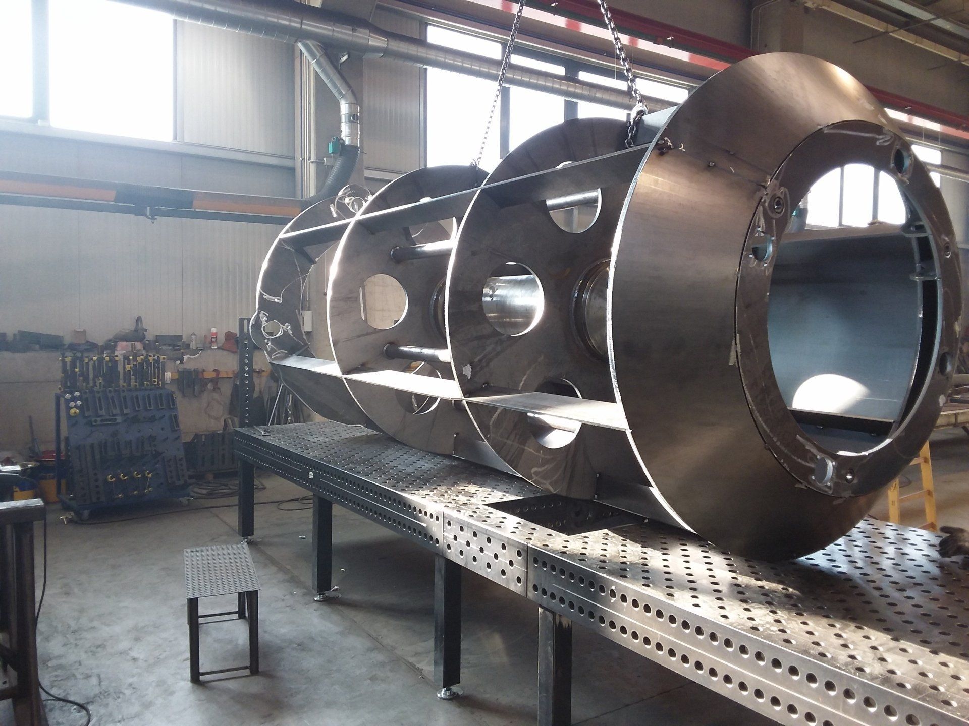 In un'officina, un grande manufatto industriale metallico di forma cilindrica è appoggiato su un banco di saldatura.