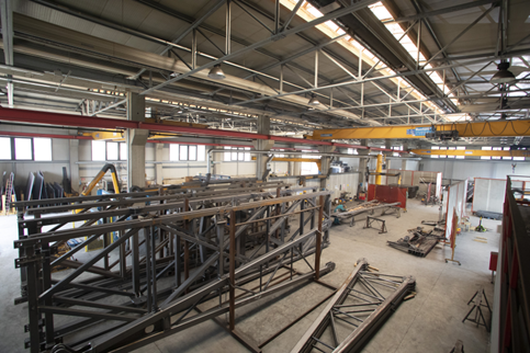 Vista dall'alto di una vasta officina industriale con grandi tralicci metallici in acciaio stoccati sul pavimento di cemento.