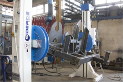 In un'officina, un posizionatore di saldatura Sideros tiene fermo un grande gruppo metallico tra due supporti rotanti blu.