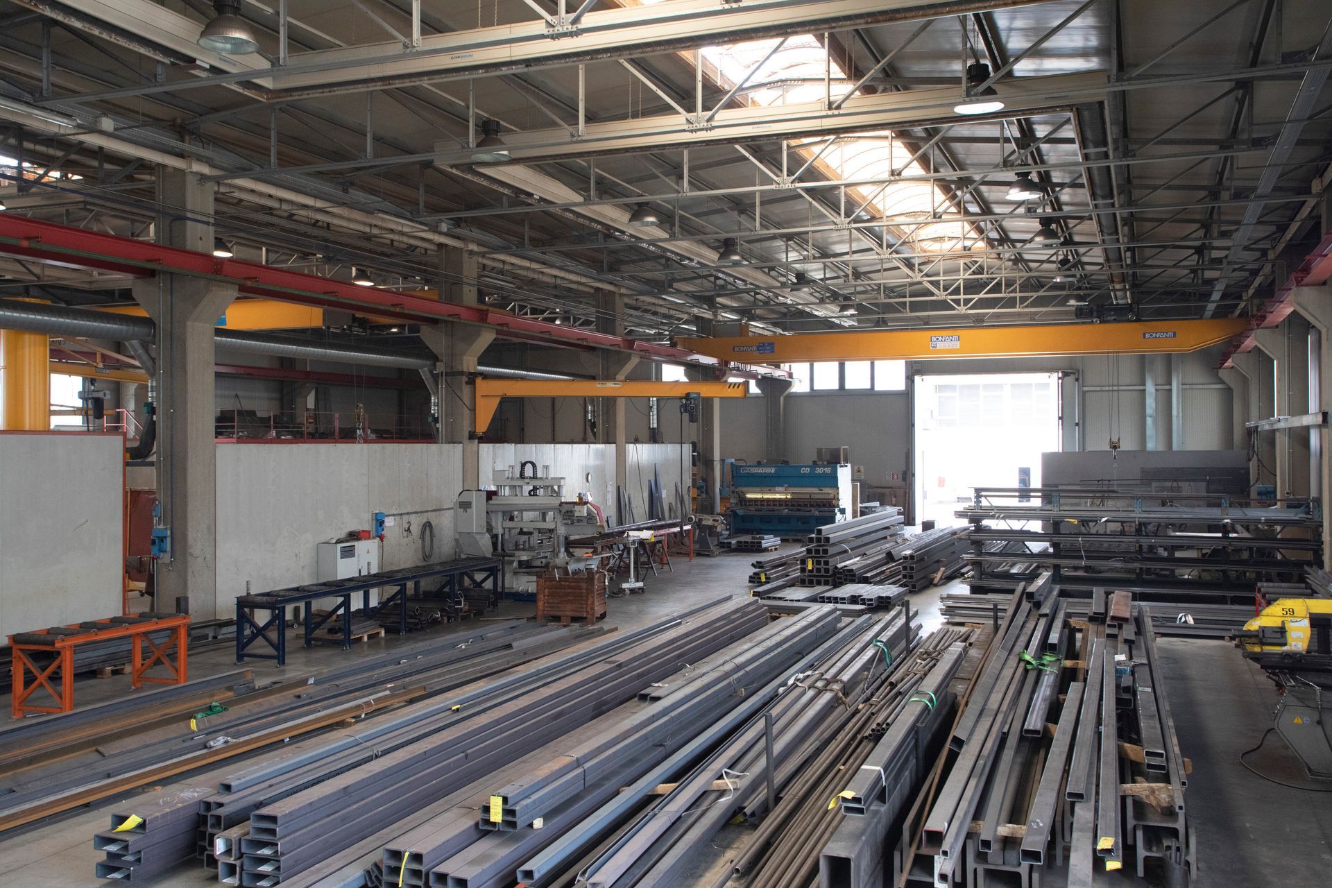 Il pavimento di un magazzino industriale è caratterizzato da pile di travi e tubi metallici, con gru a ponte gialle e soffitti alti.