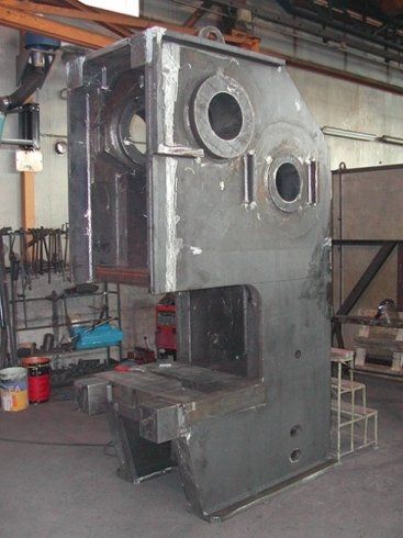 All'interno di un'officina, si trova una grande pressa industriale in metallo grigio, caratterizzata da aperture circolari e una base piatta su un pavimento in cemento.