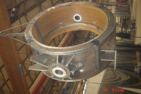 In un'officina, un grande componente industriale metallico di forma circolare è sospeso tramite catene.
