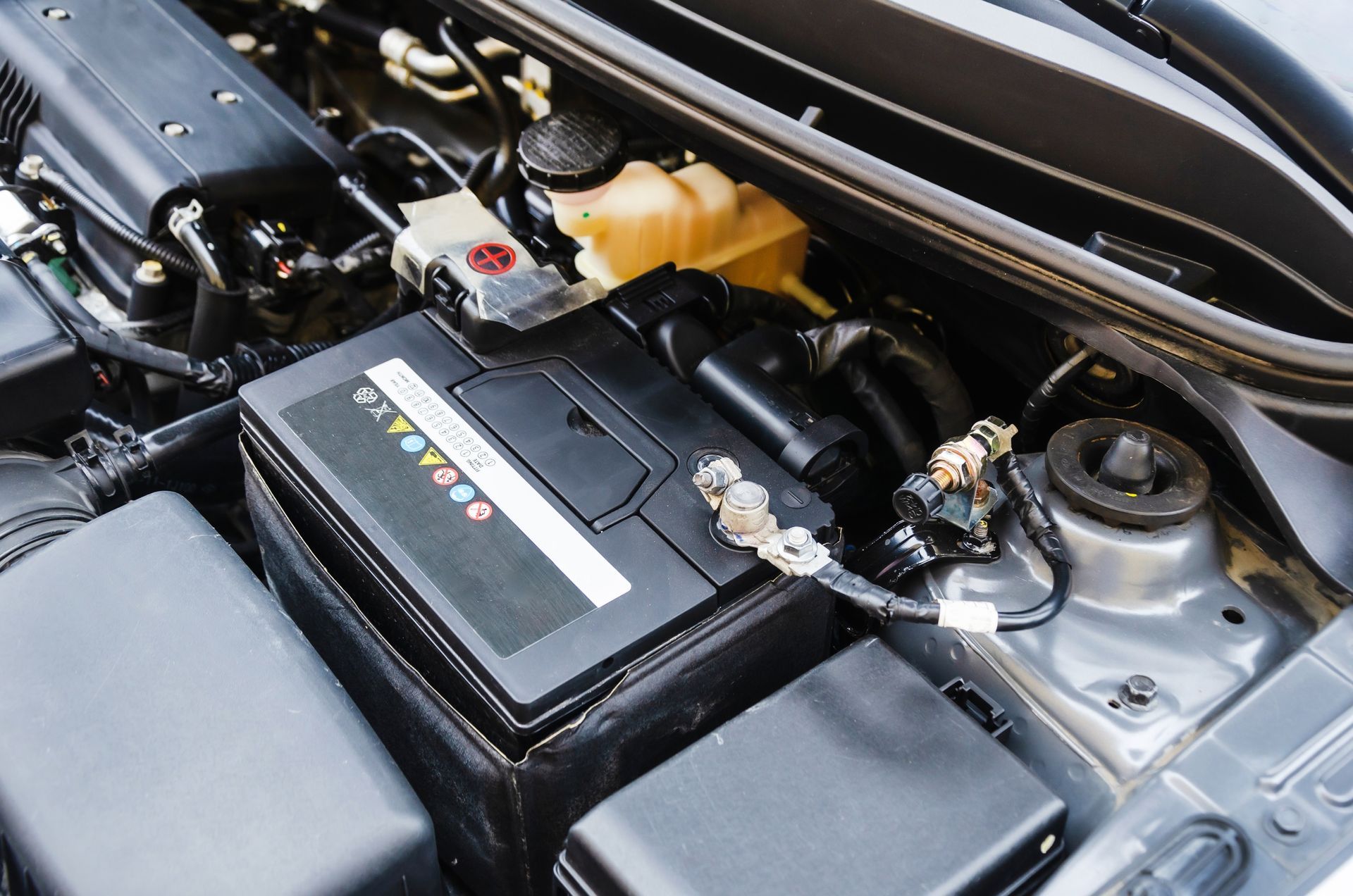 Car battery installed in a car engine bay, with cables and fluid reservoir visible.
