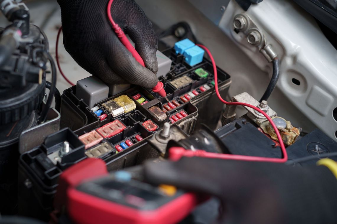 Gloved hand working on a car fuse box with red wires and exposed relays under the hood