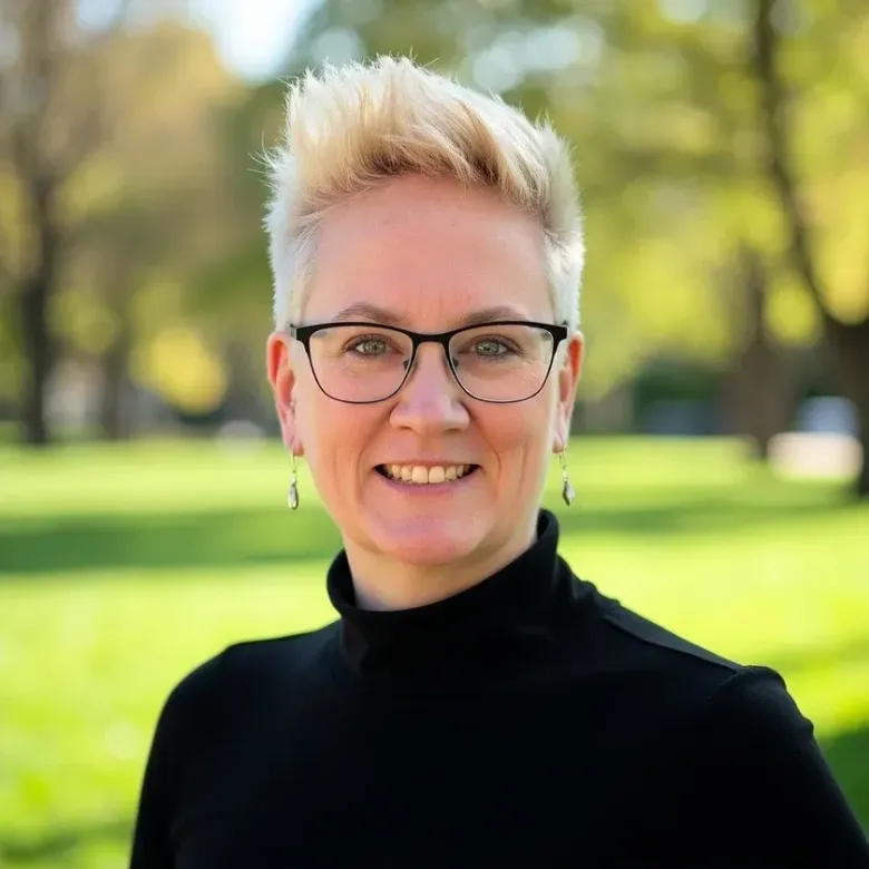 Smiling person with short blond hair and glasses outdoors in a black turtleneck on a sunny day