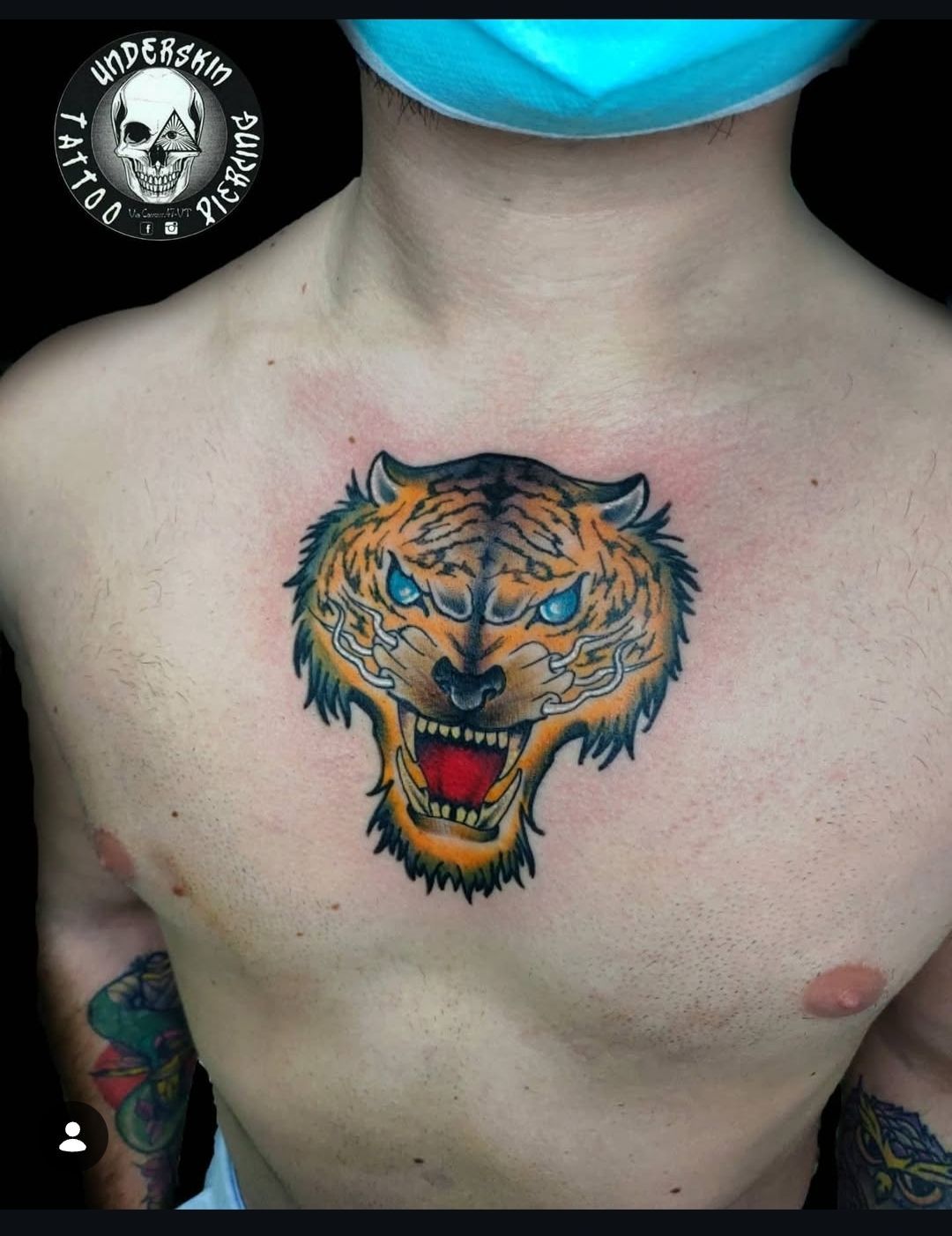 Tatuaggio sul petto raffigurante il volto di una tigre ruggente con dettagli arancioni, neri e blu su torso nudo.