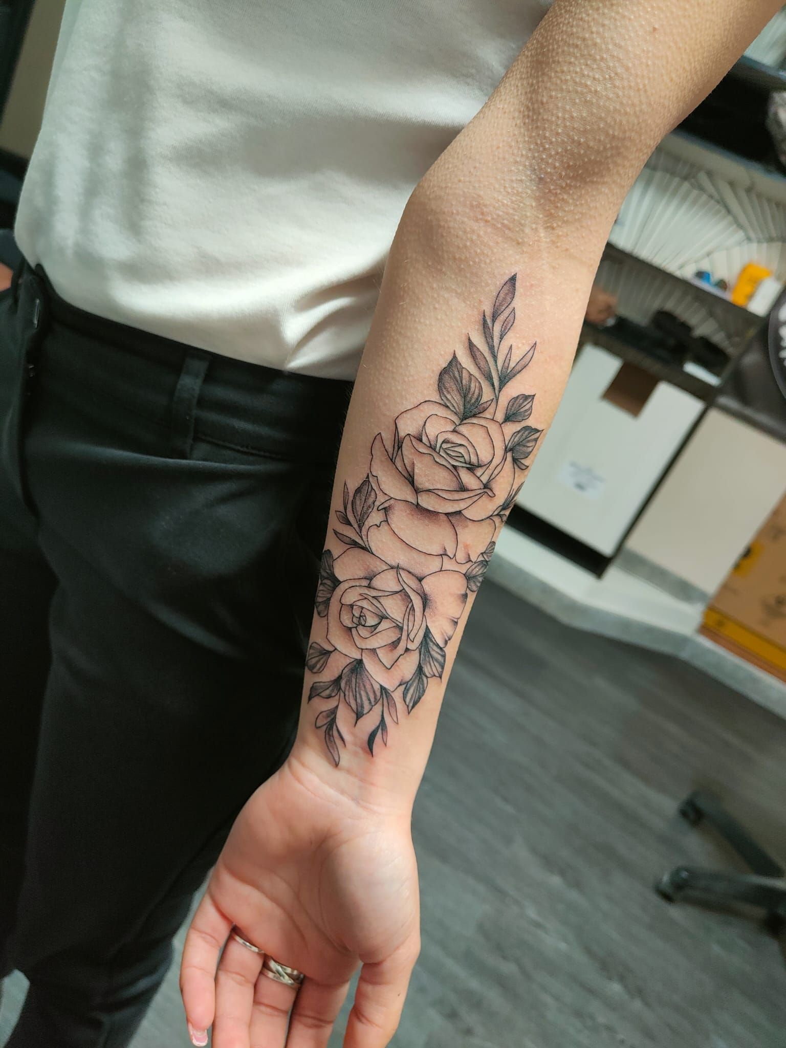 Avambraccio con tatuaggio floreale nero, mano rilassata accanto a pantaloni neri e camicia bianca in casa