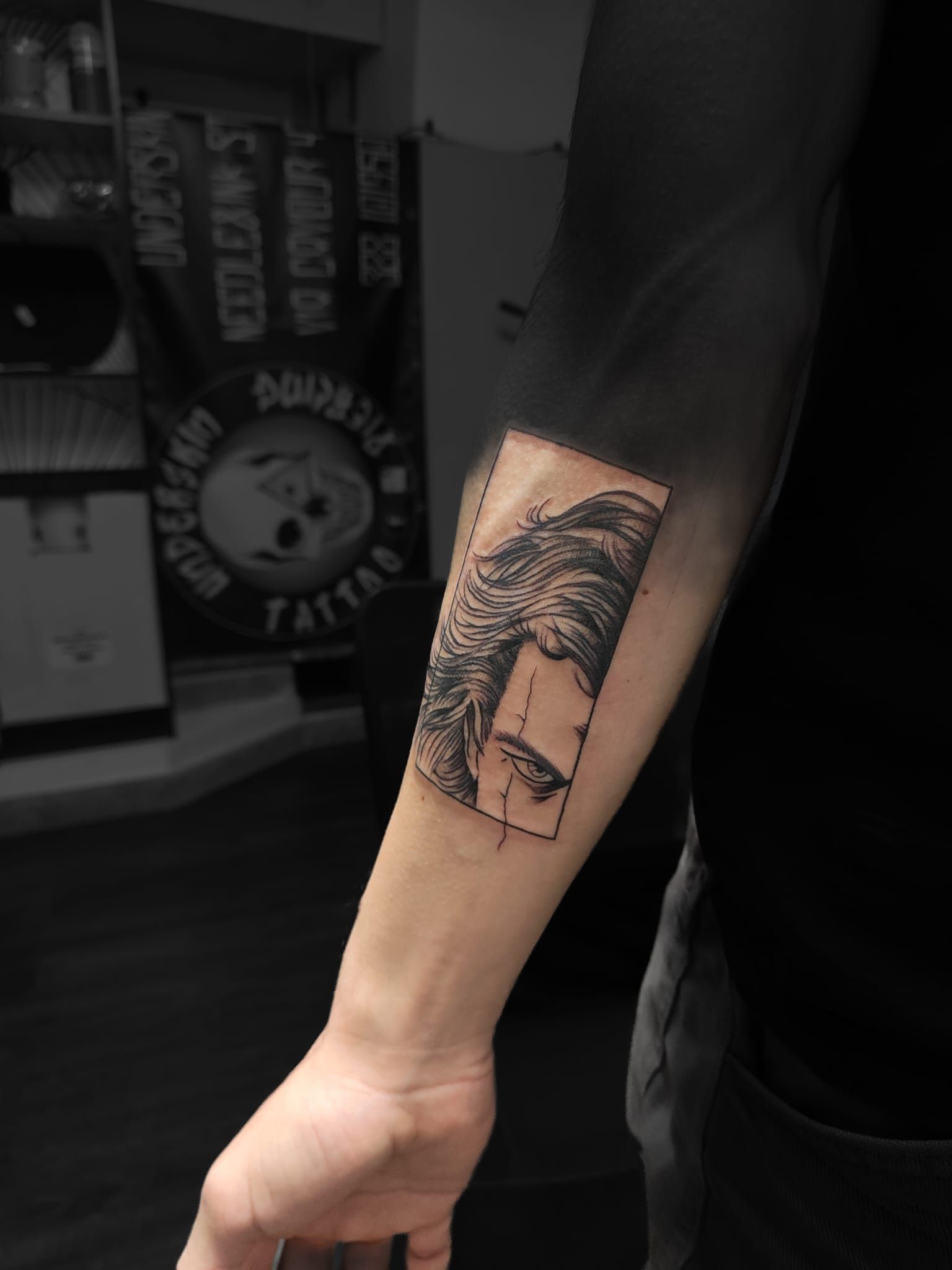 Avambraccio con tatuaggio in bianco e nero di un volto mascherato e montagne in uno studio di tatuaggi poco illuminato.