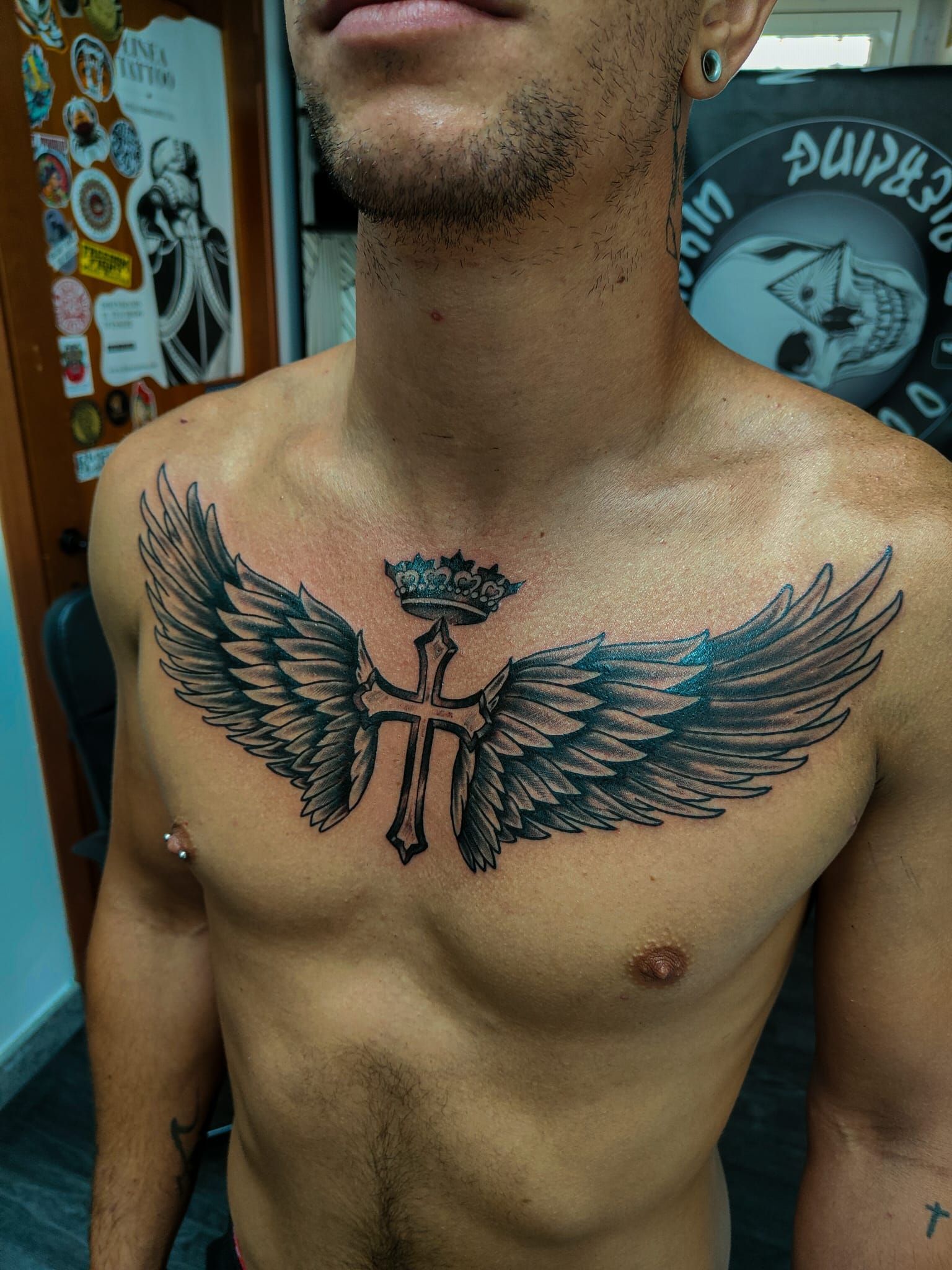 Torso nudo tatuato con un grande tatuaggio alato sul petto, in piedi davanti a muri ricoperti di graffiti.