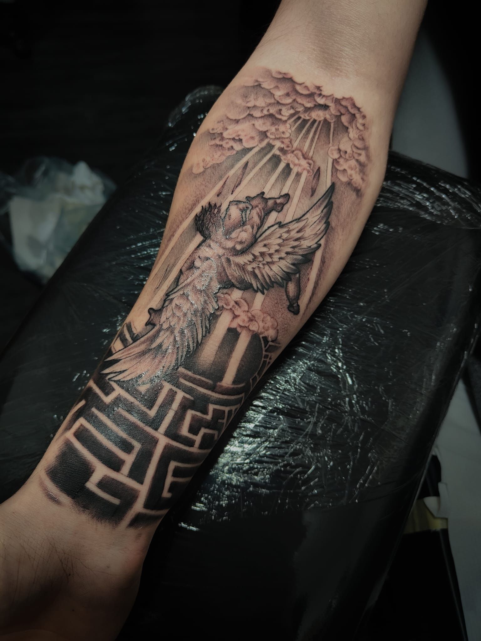 Tatuaggio sull'avambraccio raffigurante una figura alata sopra un motivo geometrico, realizzato con inchiostro nero e grigio.