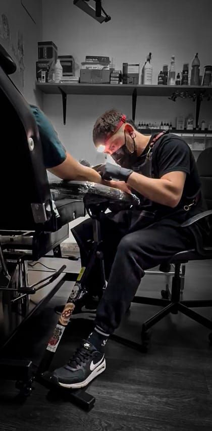 In uno studio, con scaffali pieni di materiali e mascherina, un tatuatore lavora sul braccio di un cliente.