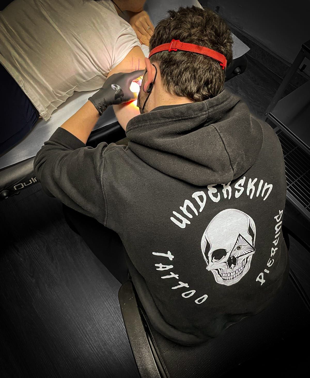 Un tatuatore con indosso una felpa nera con il logo di un teschio tatua un cliente sul braccio in uno studio.