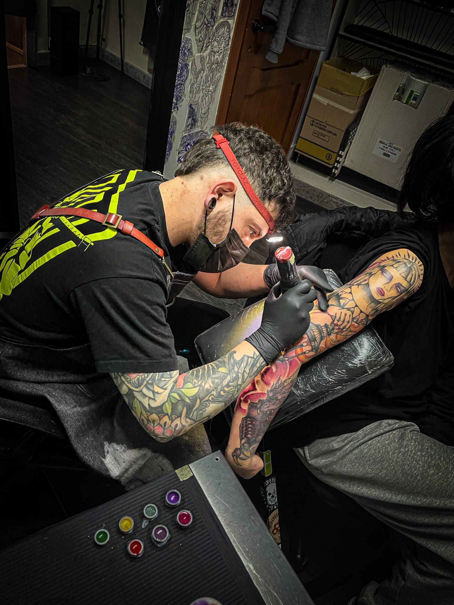 In uno studio, un tatuatore con mascherina e fascia per la testa lavora a un intricato tatuaggio sul braccio di un cliente.