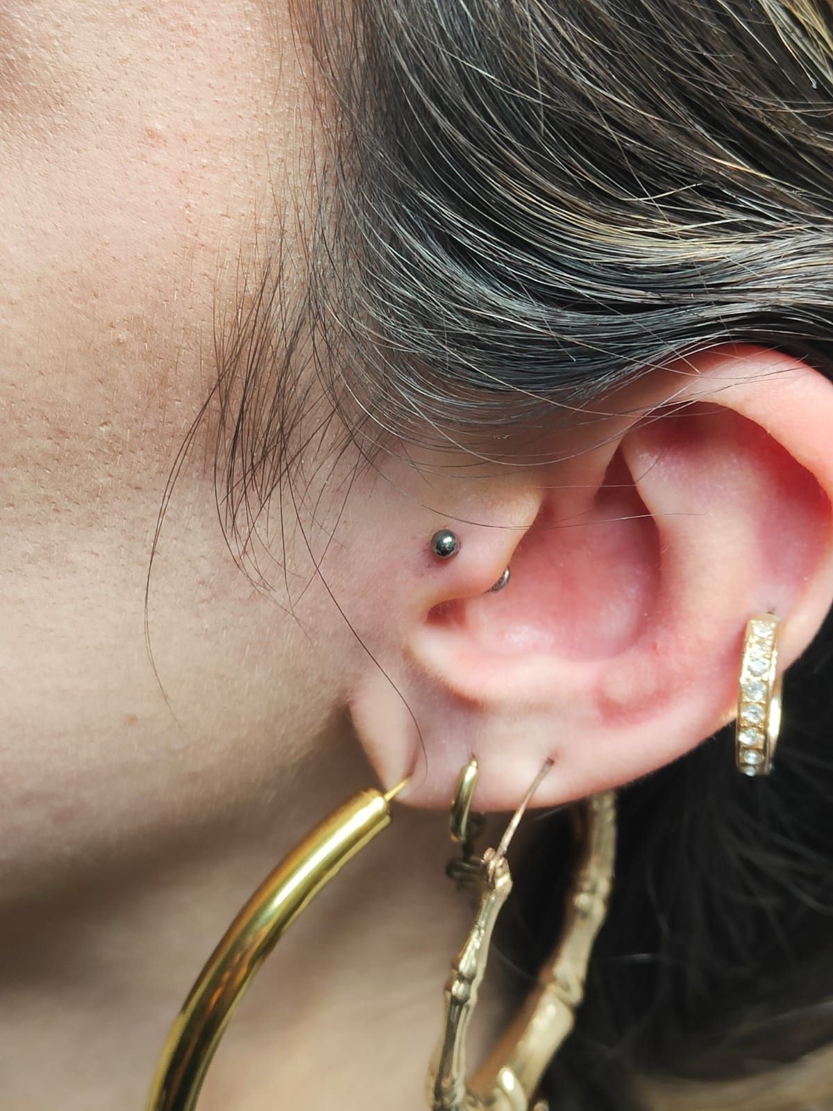 Primo piano di un orecchio con diversi orecchini e un piccolo piercing al naso, incorniciato da capelli scuri.