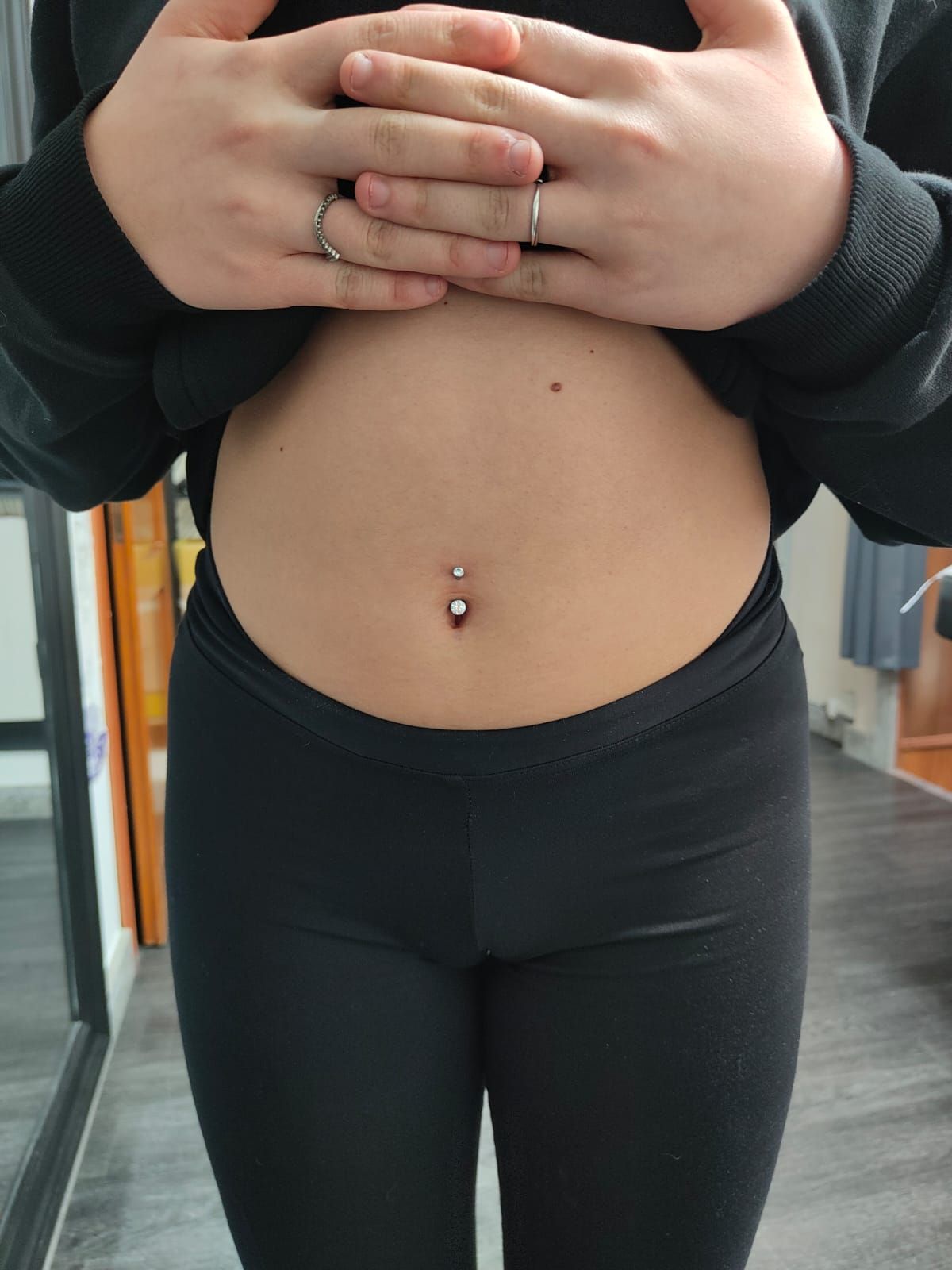 Una persona con top e leggings neri mostra il busto con un piercing all'ombelico in un ambiente interno.