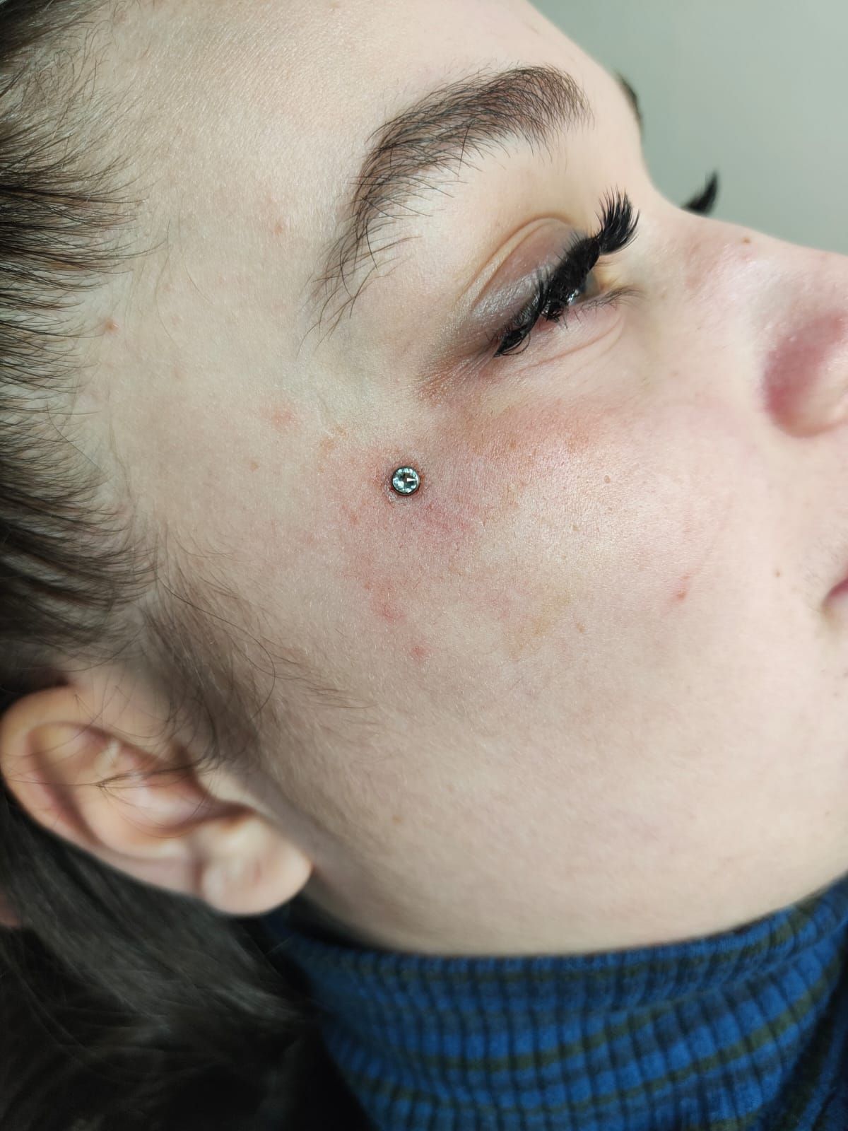 Primo piano di un viso con un piccolo piercing sotto l'occhio, che indossa un dolcevita blu.
