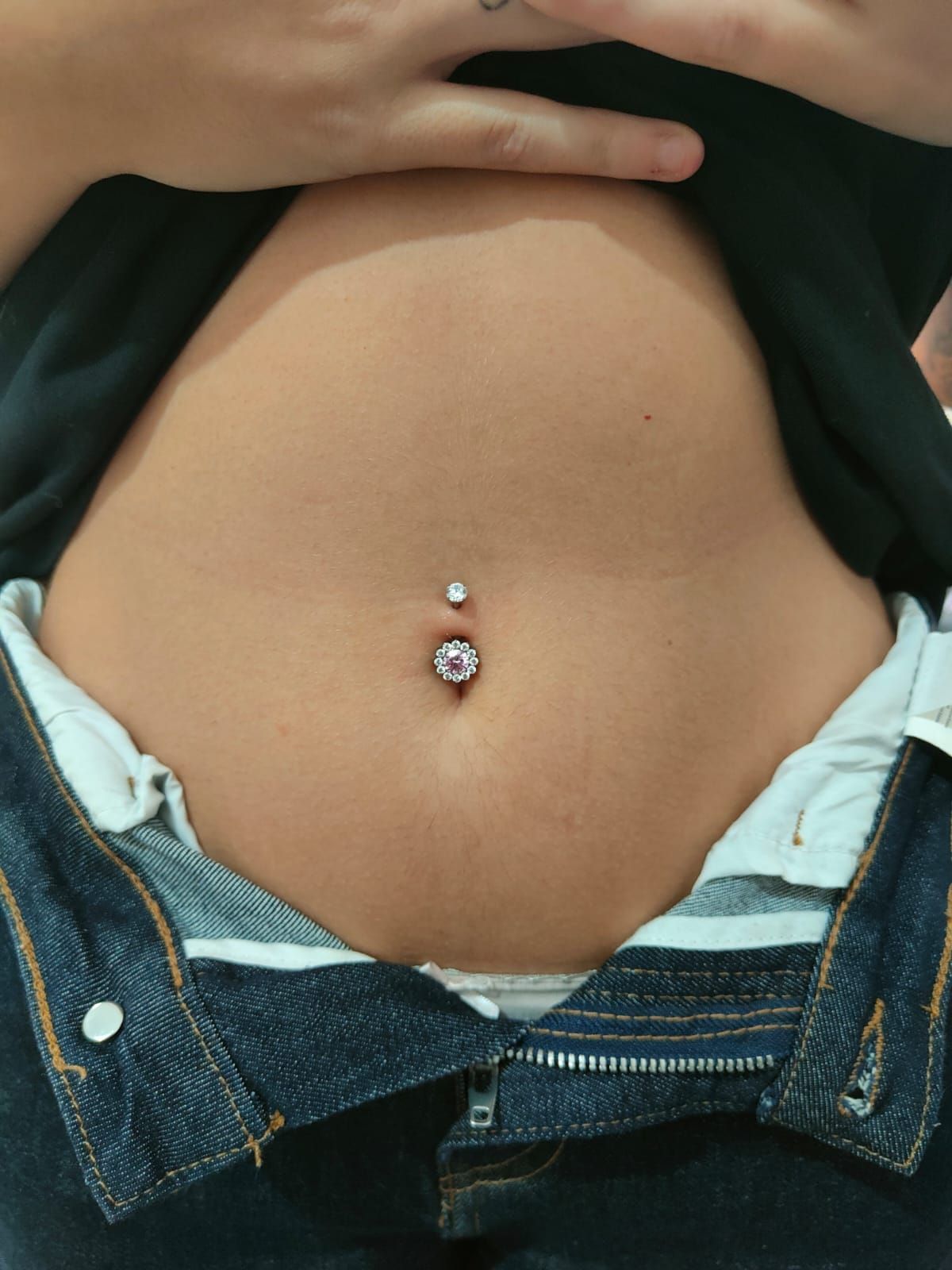 Pantaloncini corti con piercing all'ombelico, jeans blu sbottonati e una maglietta nera sollevata per mostrare la pancia