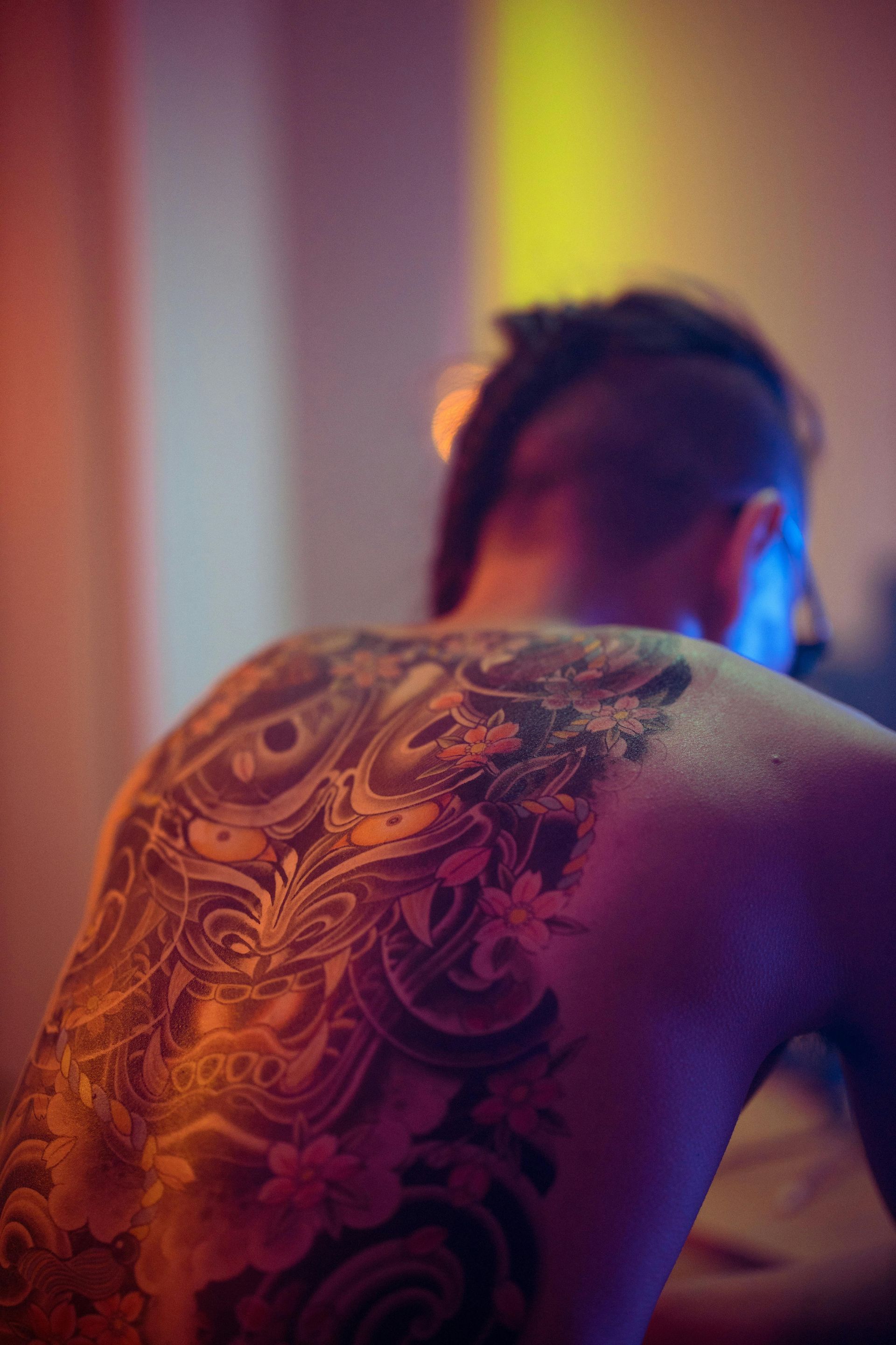 Persona con un elaborato tatuaggio sulla schiena, illuminata da luci al neon colorate, vista di spalle e leggermente sfocata.