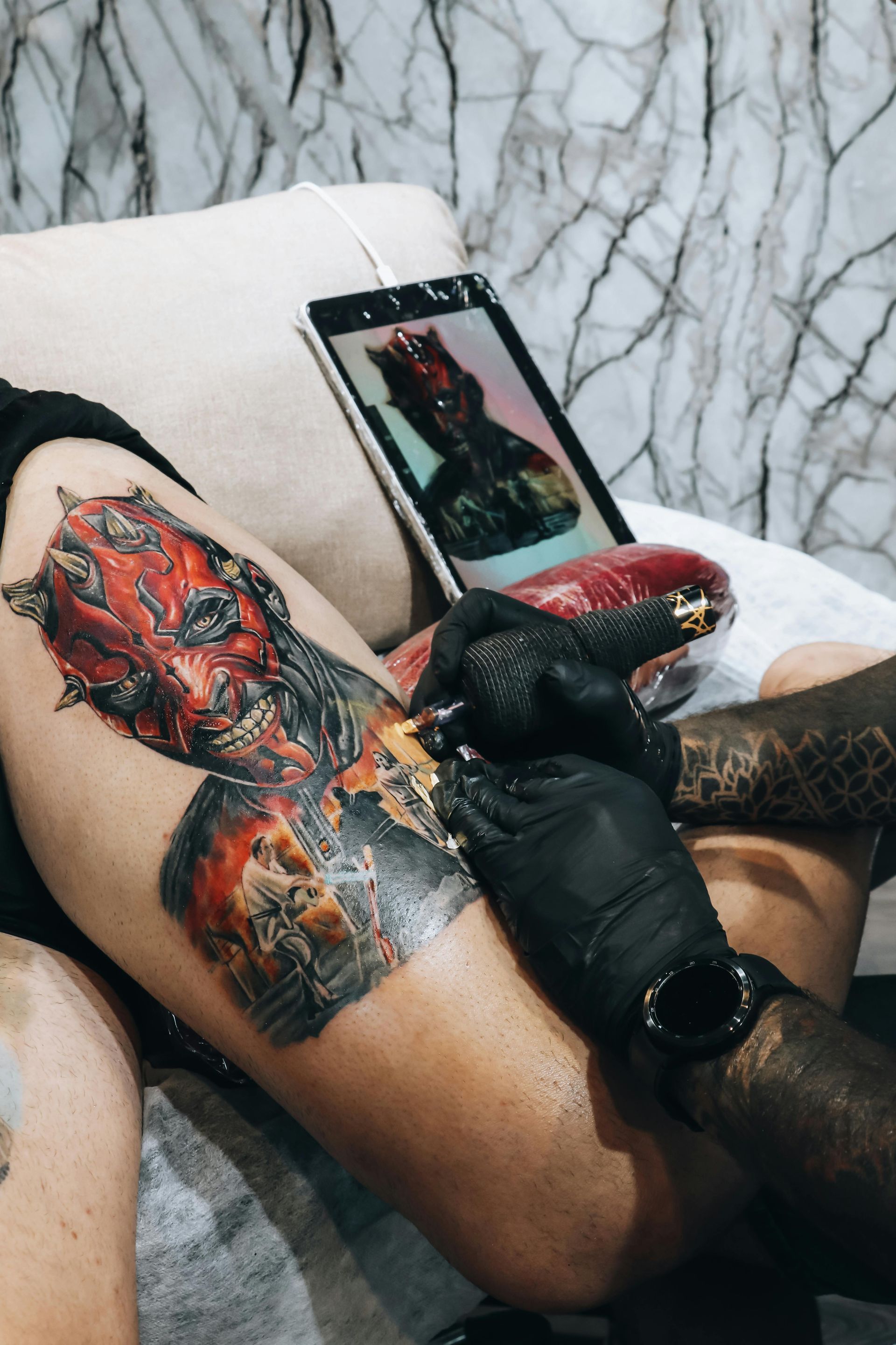 Un tatuatore esegue un tatuaggio sulla gamba con un disegno rosso e nero, utilizzando un telefono come riferimento.