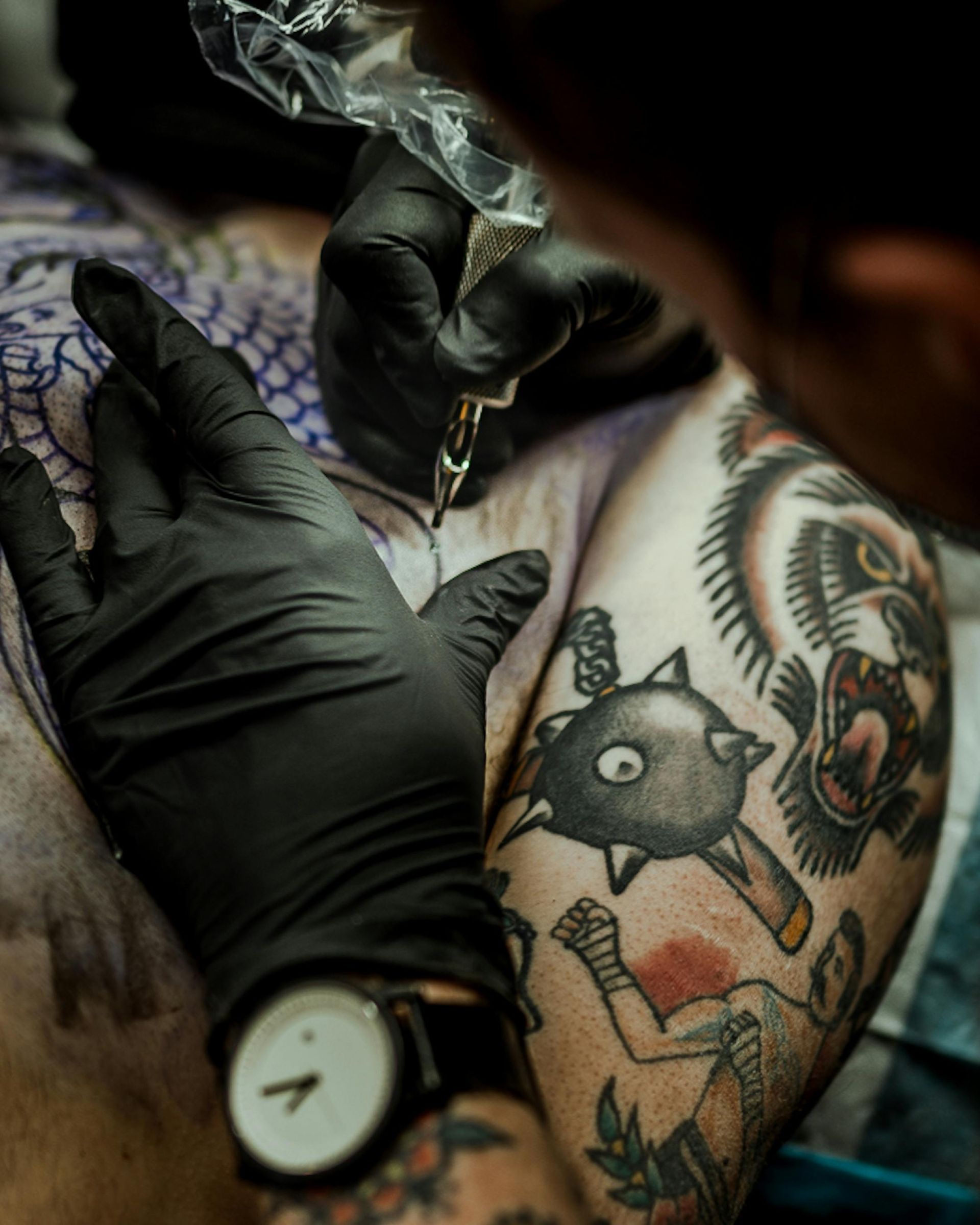 Tatuatore con guanti neri che tatua la parte superiore del braccio di una persona con una macchinetta.