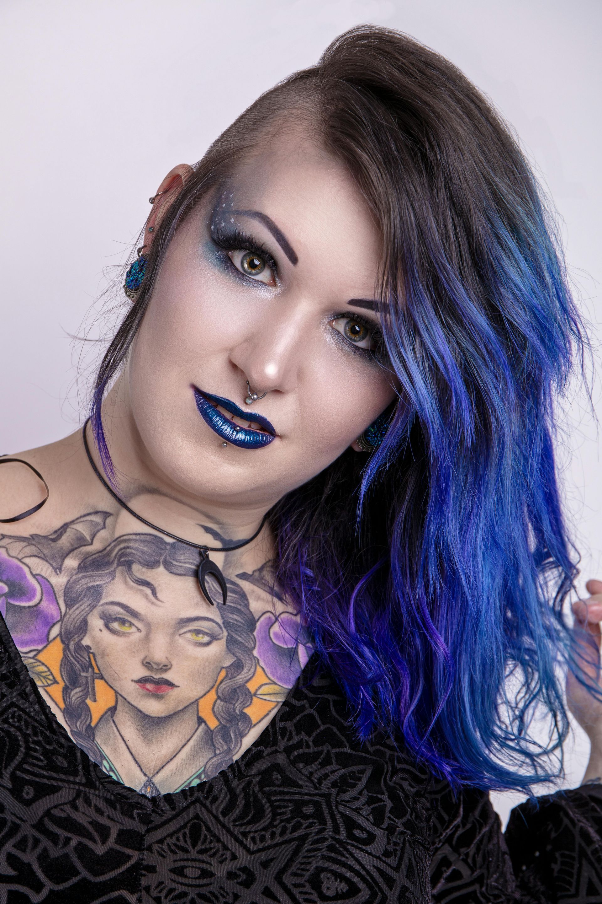 Una persona con i capelli striati di blu, il rossetto scuro e un elaborato tatuaggio sul petto davanti a uno sfondo bianco.
