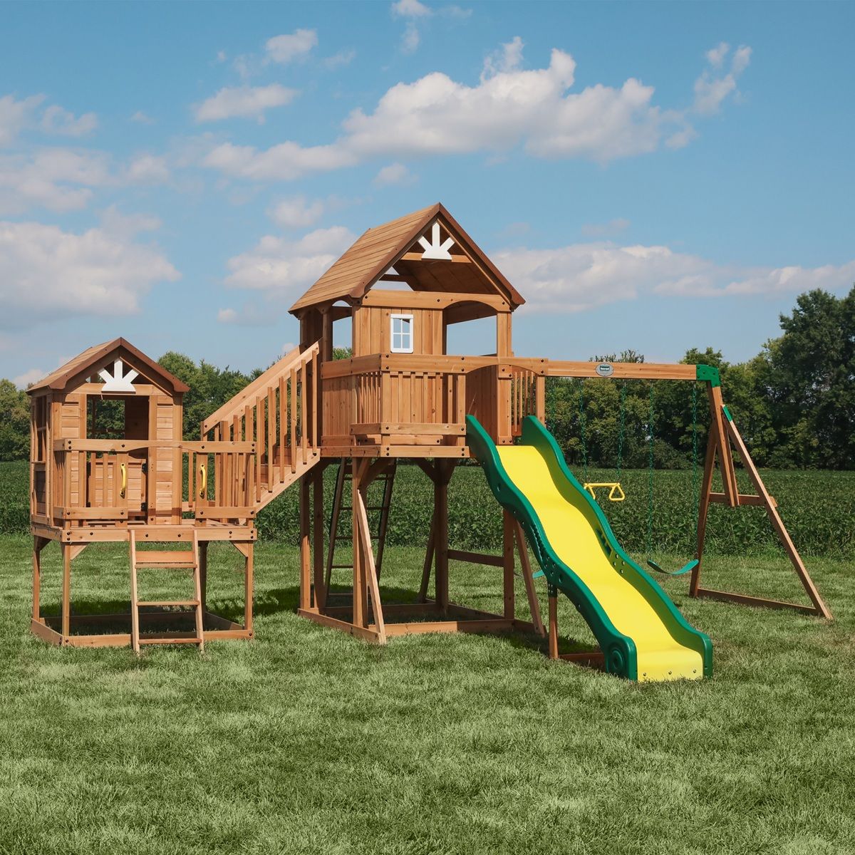 Wooden backyard playground with towers, slide, and swing set on a grassy lawn under a blue sky