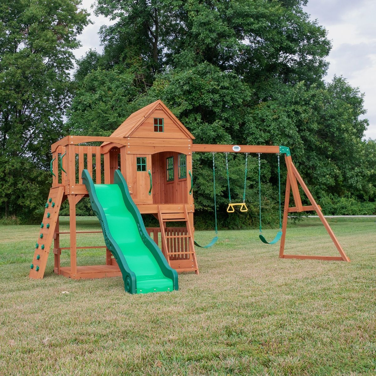 Wooden backyard playset with green slide, swing set, and tower on a grassy lawn
