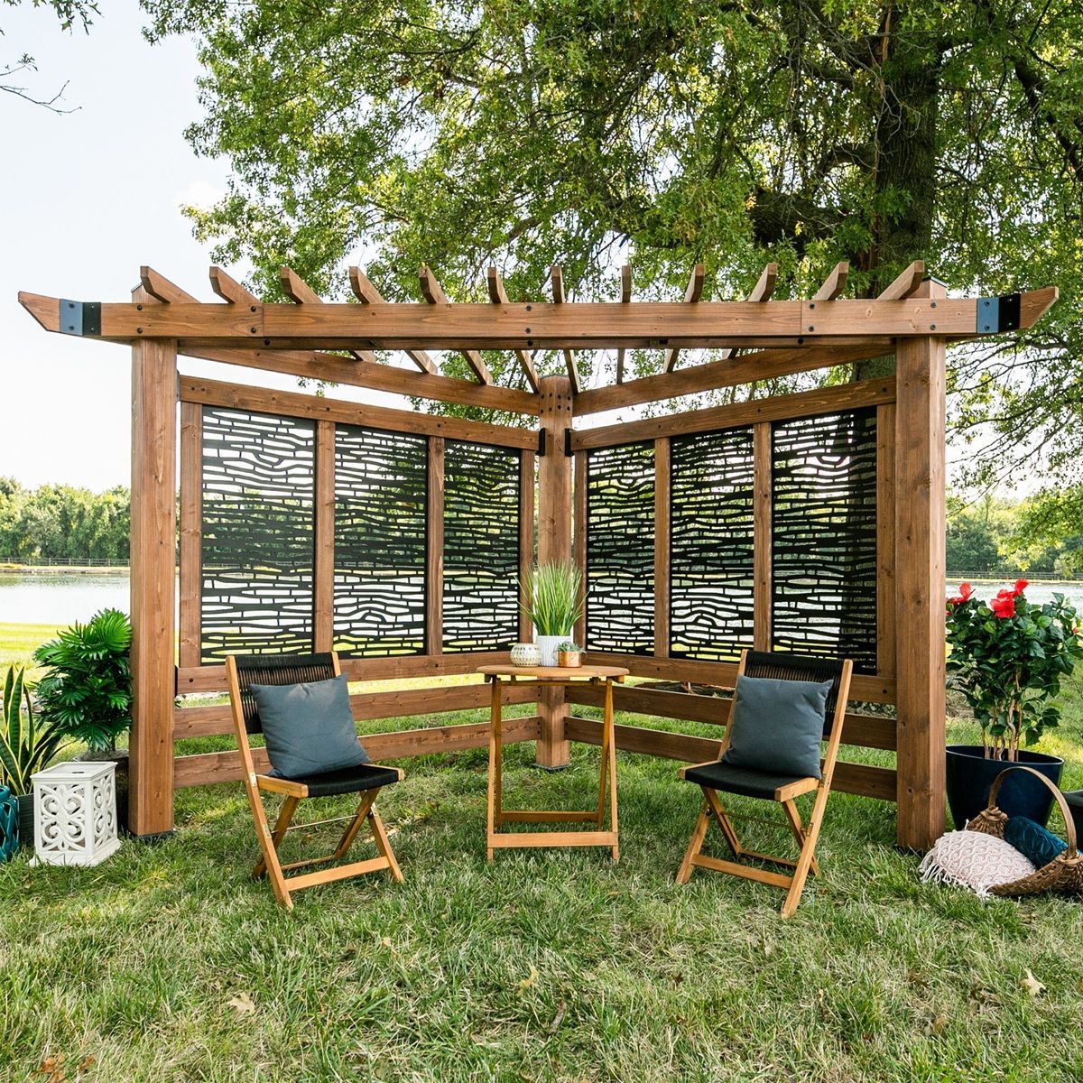 Wooden outdoor gazebo with woven walls, two chairs, and a small table in a grassy garden by a lake