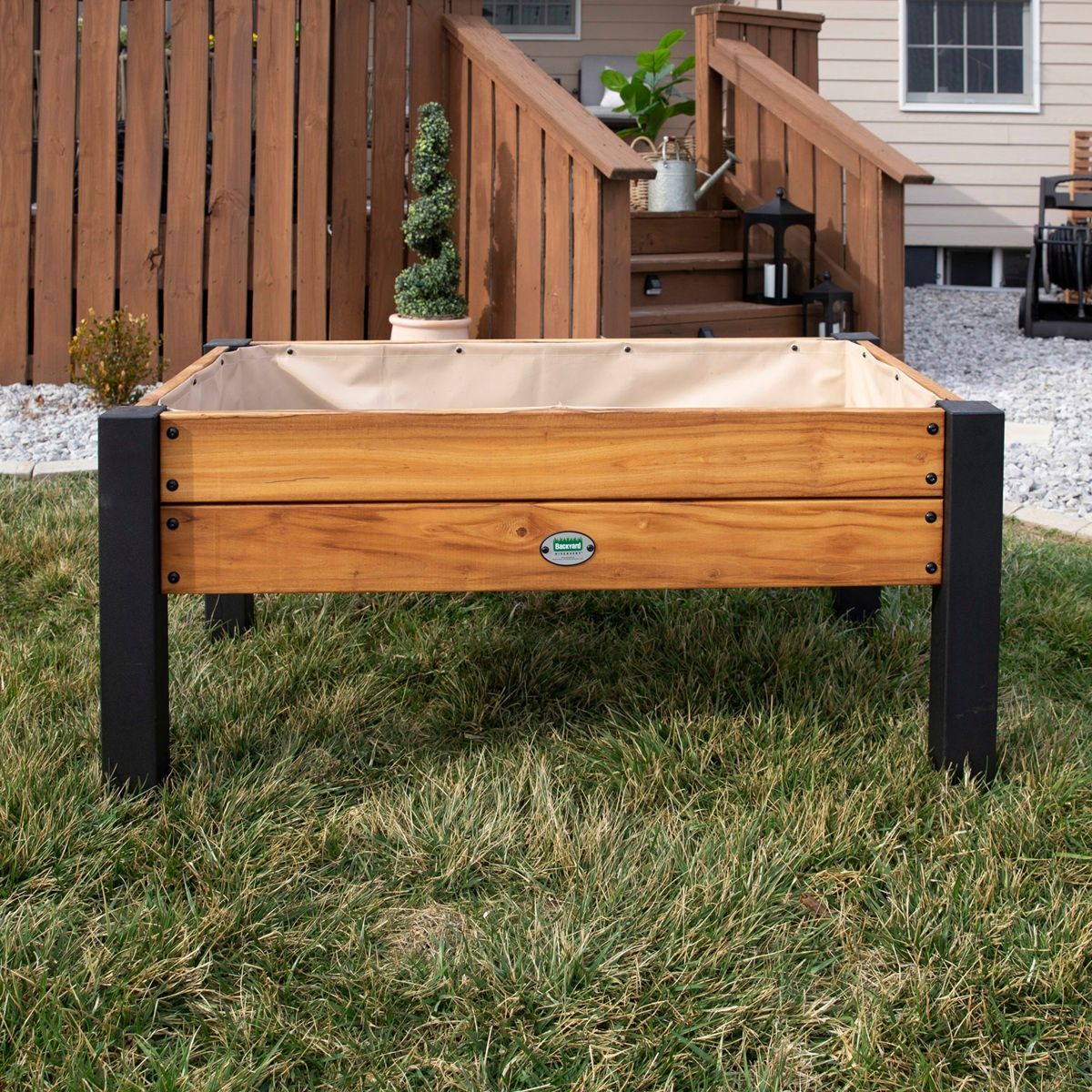 Wooden raised garden bed with tan cover on black legs in a grassy yard near a house deck