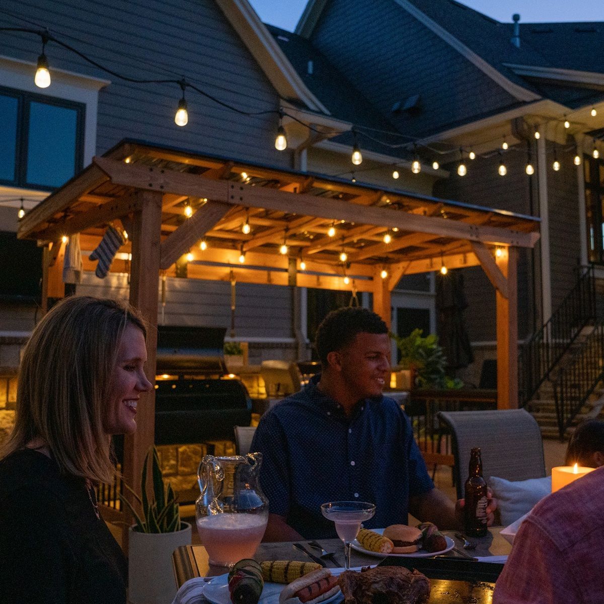 People dining outdoors under string lights at dusk in a backyard patio with a wooden pergola.