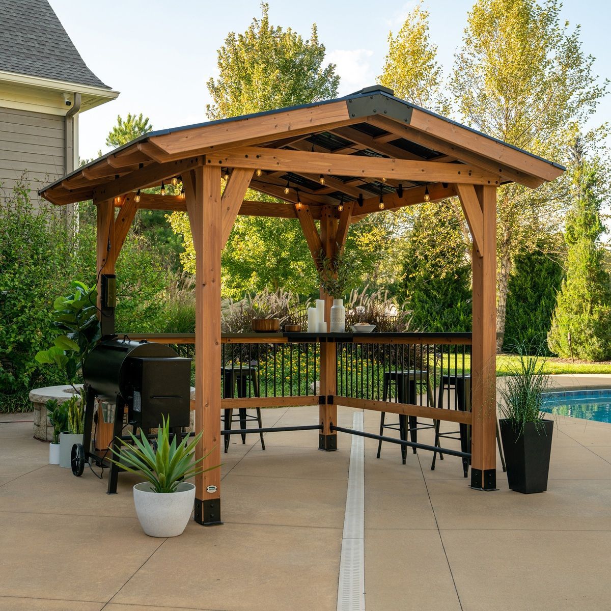 Wooden gazebo with bar seating on a patio beside a swimming pool, surrounded by potted plants and trees