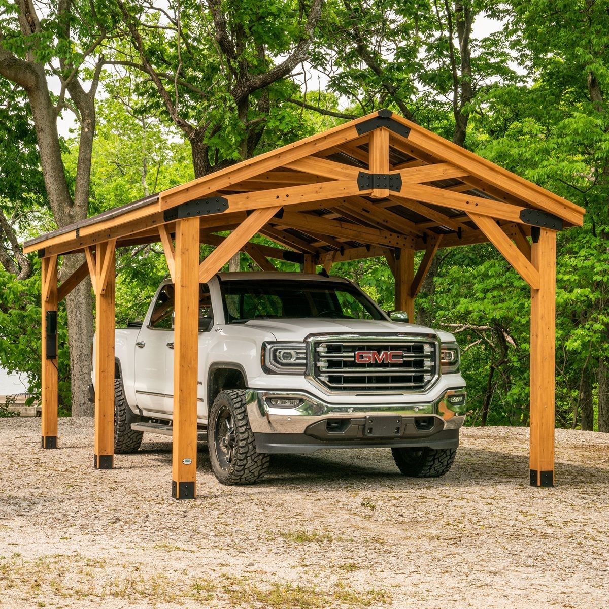 White GMC truck parked under a wooden carport in a gravel yard surrounded by green trees.