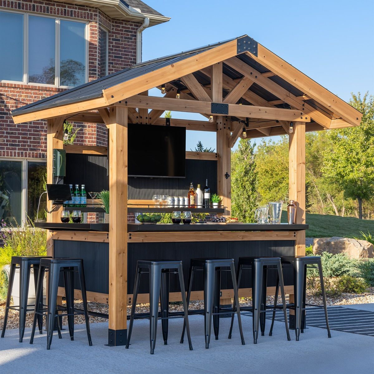 Wooden outdoor bar with black stools and a gabled roof beside a brick house